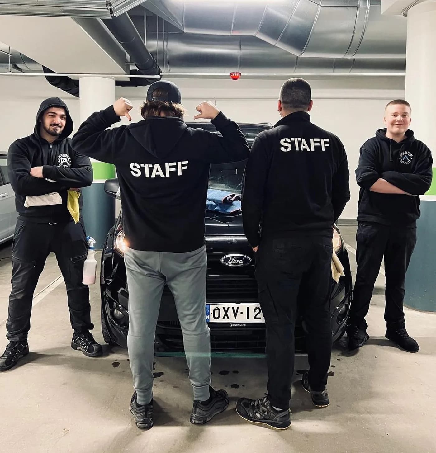 Premium Autopesun neljä työntekijää mustissa STAFF-huppareissa auton edessä parkkihallissa